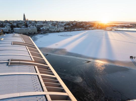 The roof of City Hall in the foreground, the frozen Tjörnin pond, the sun rising in the distance. Hallgrímskirkja church and Þingholt in the background.