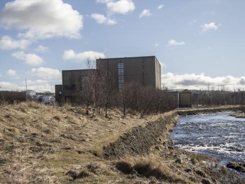 Toppstöðin í Elliðaárdal, gul sina og Elliðaá í forgrunni, blár himinn með nokkrum skýjum í bakgrunni