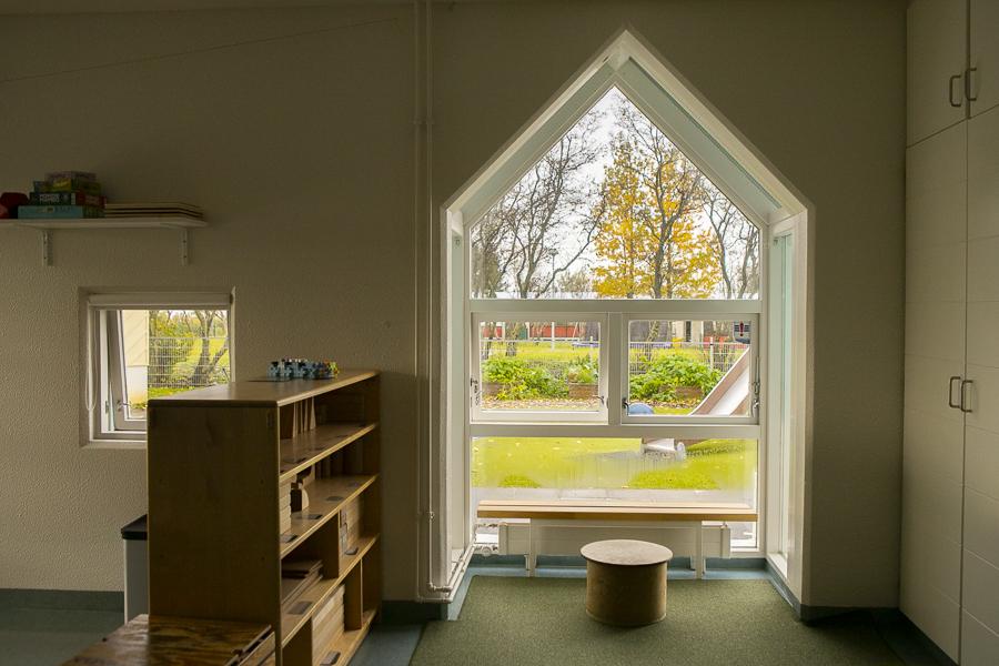 Séð út um glugga á leikskólanum Sólborg. Inni er hilla og lítill stóll en úti rennibraut og önnur leiktæki.