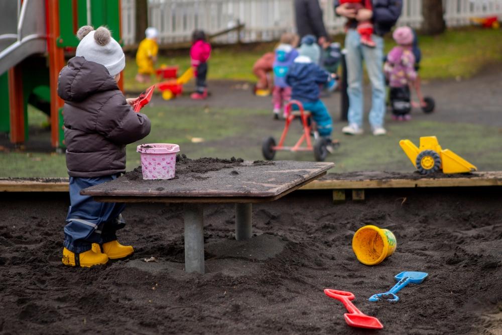 Barn að leik með skóflu og fötu í sandkassa á leikskólanum Miðborg, Barónsstíg. Fleiri börn í baksýn.