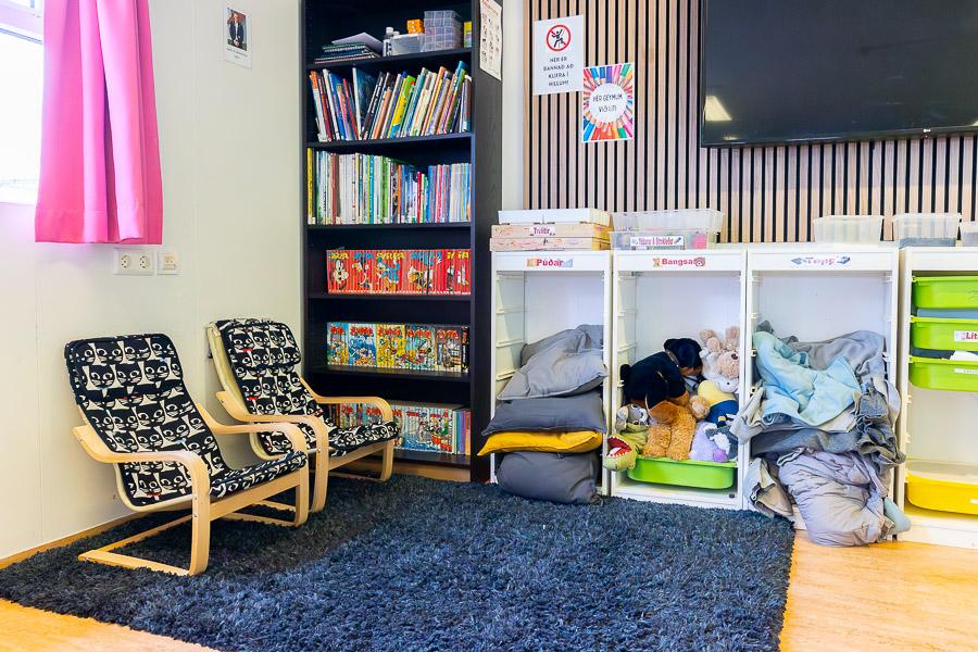 Herbergi með stólum, bókahillu með syrpum og bókum, hillur með púðum og leikföngum. Blá motta á gólfi. 