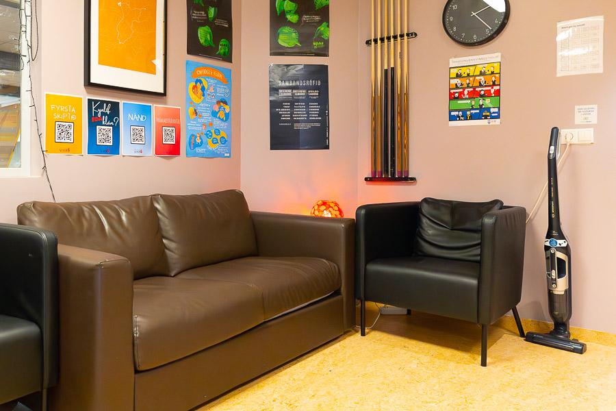 A small brown couch and black chairs. A poster, a clock, and pool cues on a light pink wall. A vacuum cleaner to the side. 