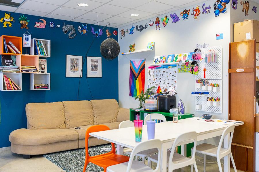 Sofa, table and chairs. Blue wall decorated with shelves and pictures. LGBT+ flag and beaded artwork displayed on the wall.
