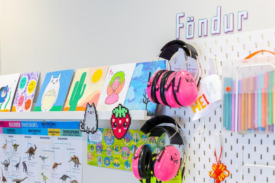 Shelf displaying pictures. Wall hooks holding pink headphones and markers. Above the hooks a sign reads "Crafts" in Icelandic. 