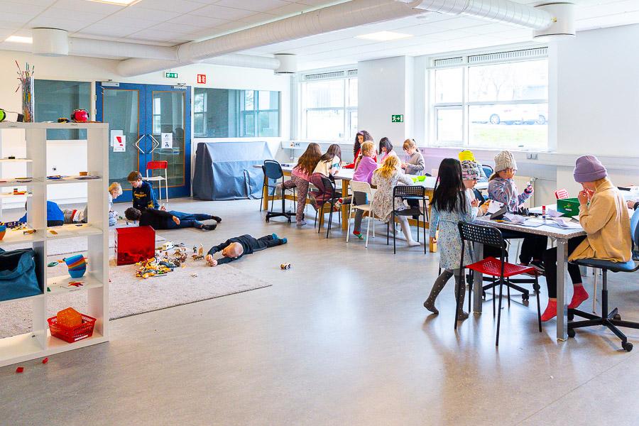Börn sitja við tvö borð að föndra. Önnur börn liggja á gólfi að leika sér með dýr og fleira. Hvít hilla stendur til hliðar.