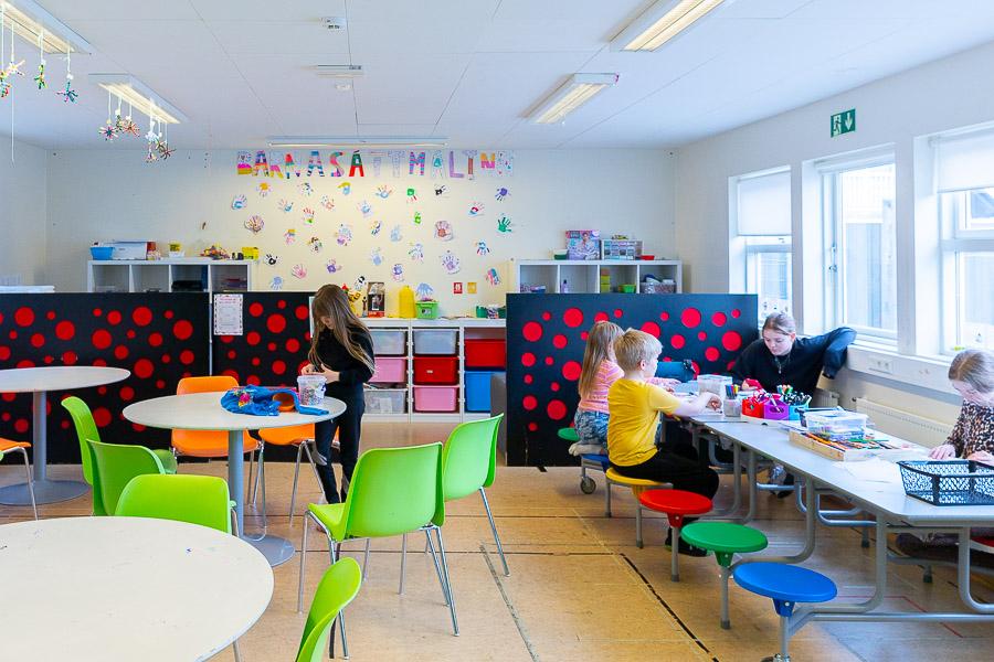 Salur með borðum og stólum. Börn sitja á marglita kollum að föndra. Í baksýn eru skilrúm með hringjamynstri. Á vegg stendur Barnasáttmáli.