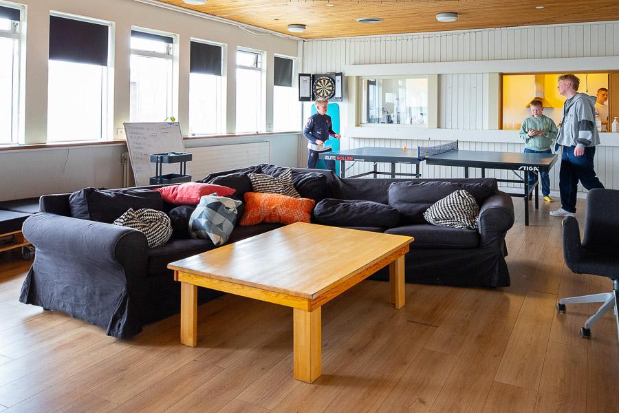 Herbergi með hornsófa, borði og borðtennisborði. Unglingar að spila borðtennis. 