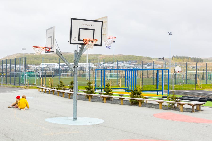 A basketball court with children sitting on the ground. A fenced soccer field in the background.