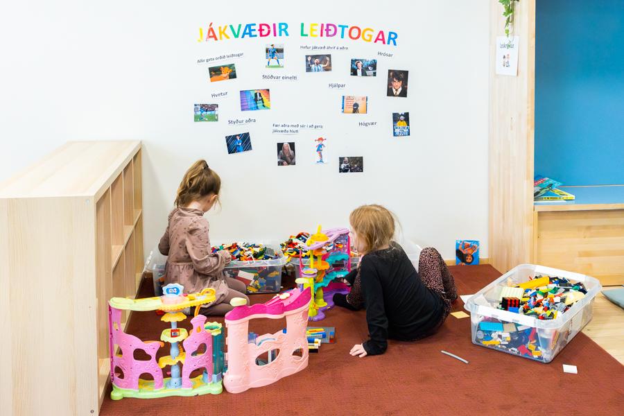 Children sit on the floor playing with Lego blocks. To the side is a shelf with wooden blocks, and on the wall behind it reads “Jákvæðir leiðtogar” (Positive Leaders). 
