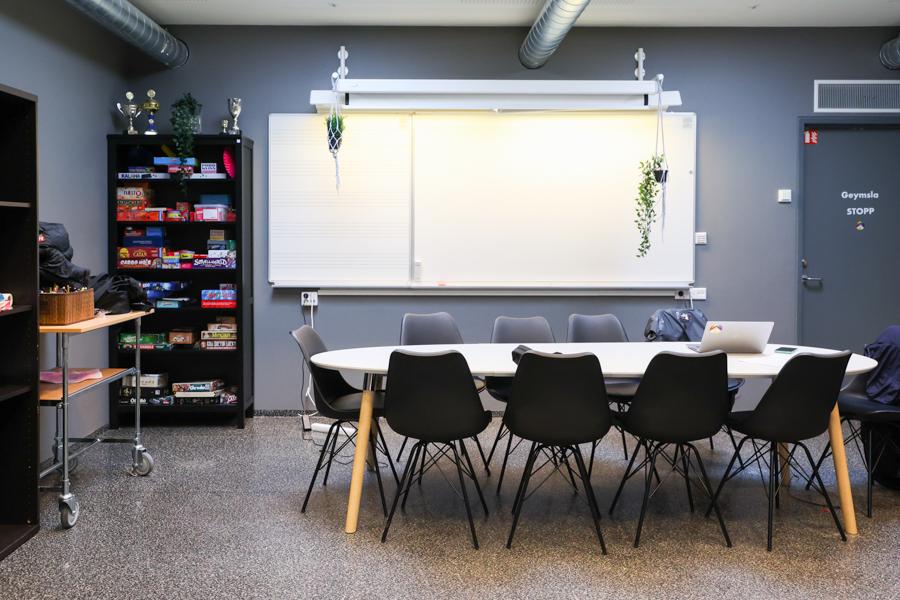 Grámálað herbergi með borði, stólum og hillu með borðspilum. 