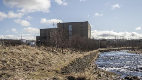 Toppstöðin í Elliðaárdal, gul sina og Elliðaá í forgrunni, blár himinn með nokkrum skýjum í bakgrunni
